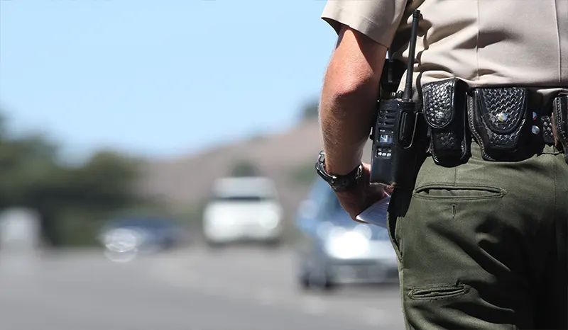 A low view of a police officer focused on his utility belt. Cars are blurred in the distance.